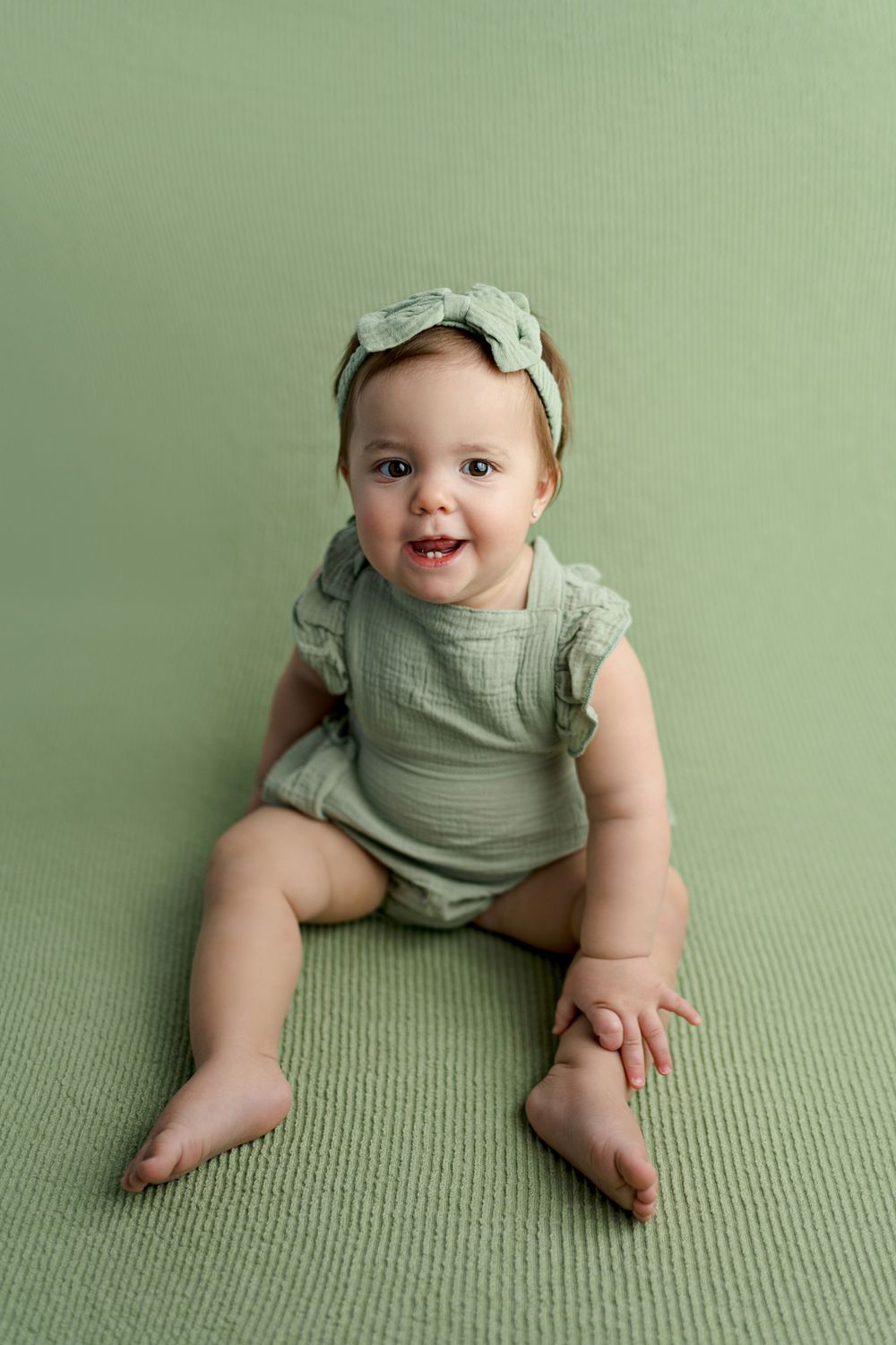 A smiling baby in sage green romper lies on matching green textured backdrop during professional photo session.