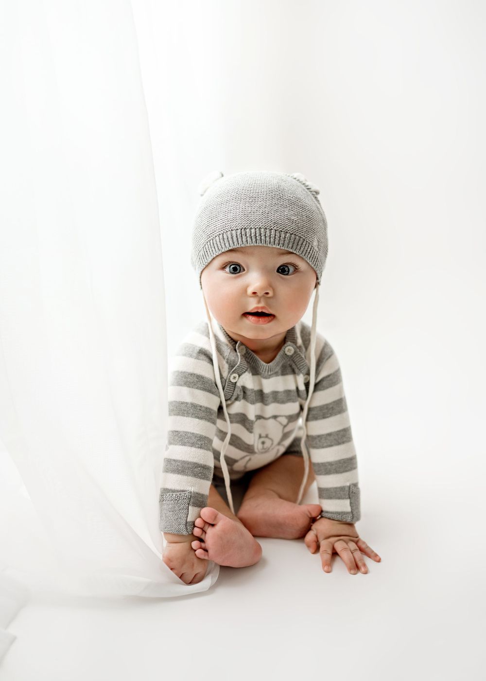 A baby wearing a gray knit hat and striped sweater sits on a white background for a portrait session.