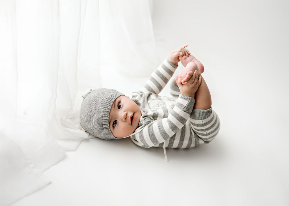 Baby wearing grey striped outfit and beanie lies on white backdrop while holding feet during minimalist photo session.