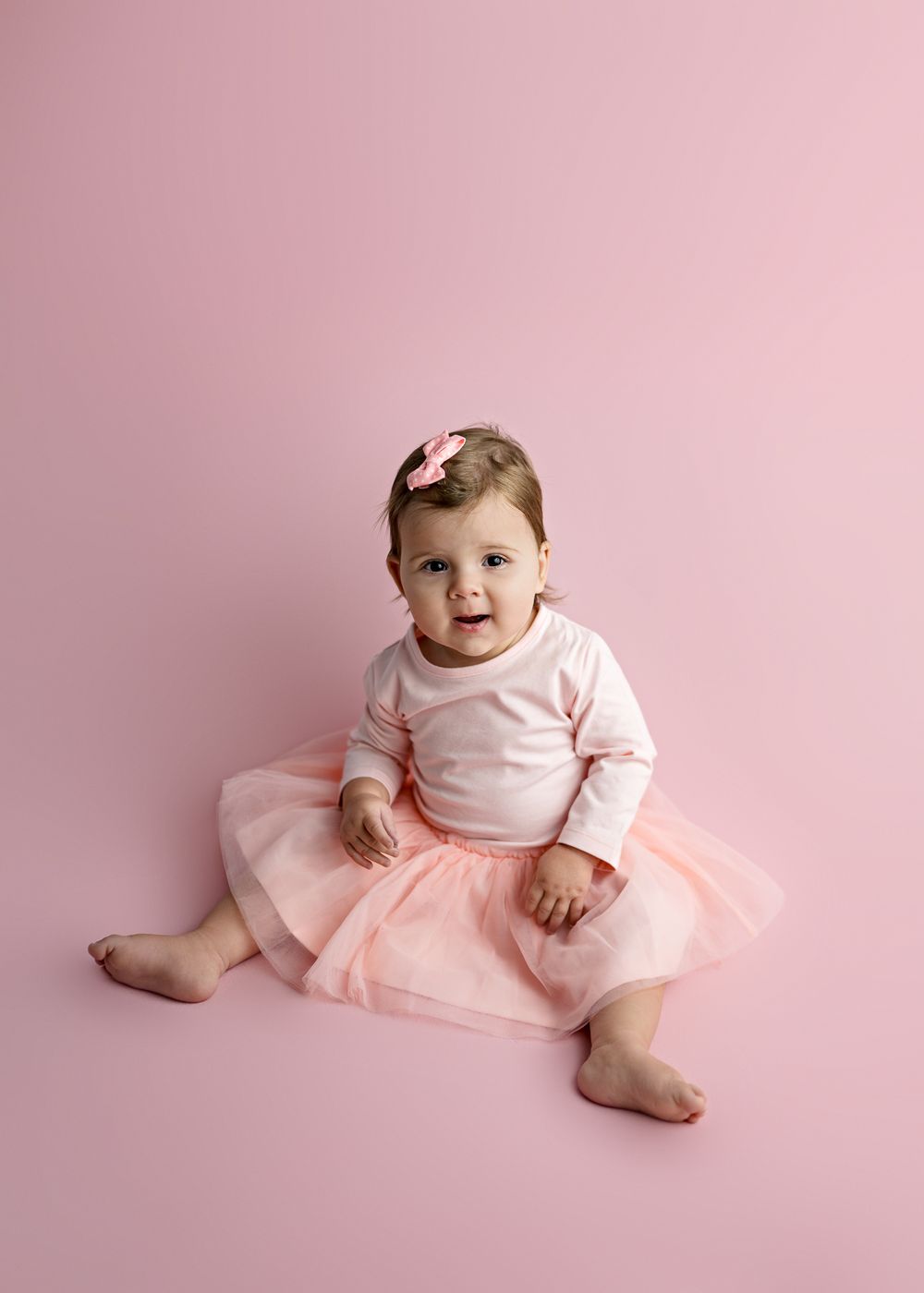 Baby wearing pink tutu dress sitting on pink background with matching hair bow.