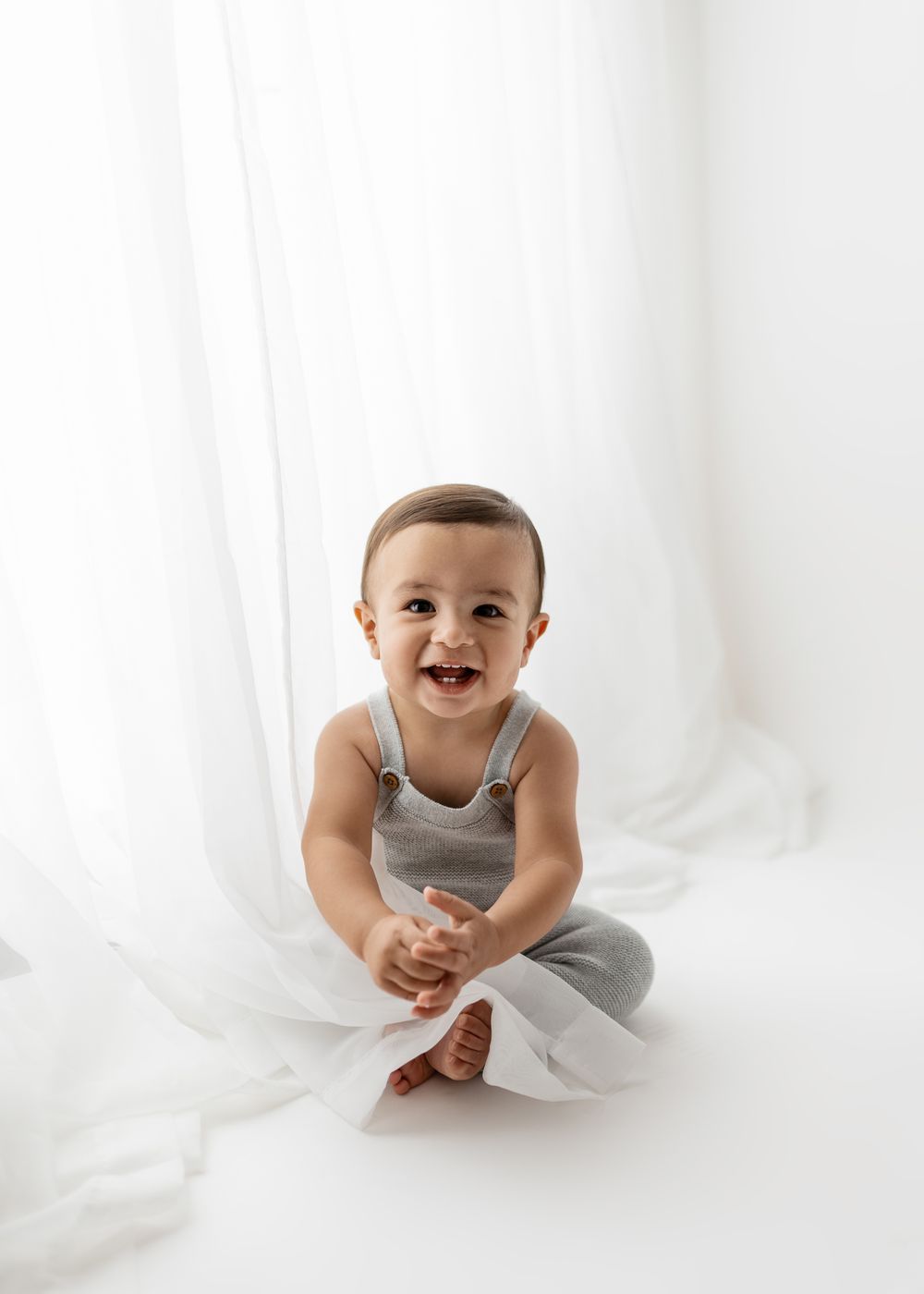 A baby in a white and gray outfit sits and smiles on a white backdrop during a studio photoshoot.