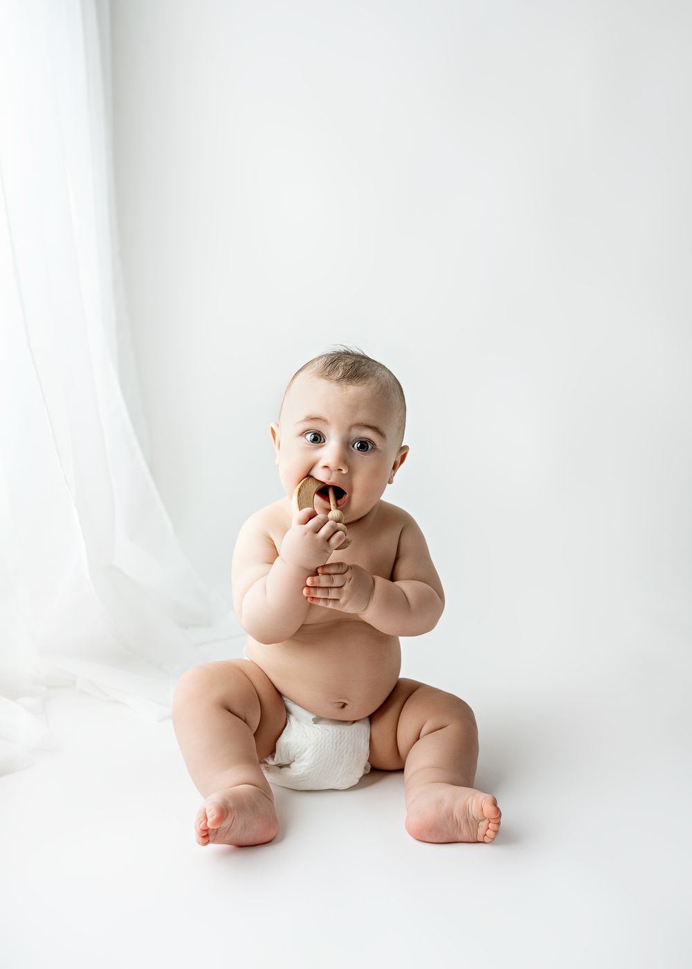 Baby wearing white diaper chewing on a cookie while sitting on white background.