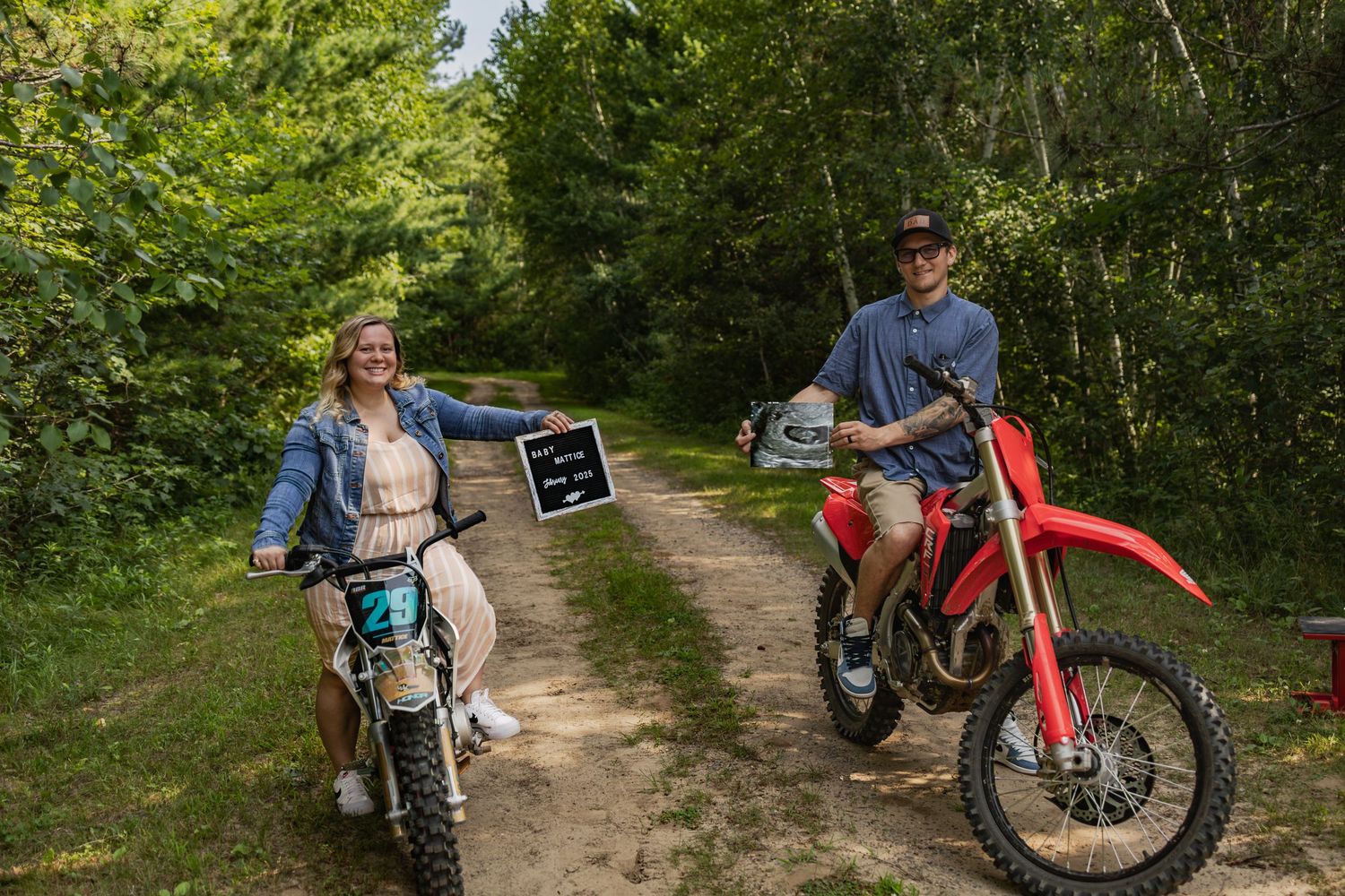 Couple posing on dirt bikes on a forest trail, holding a sign and helmet, surrounded by lush green trees.