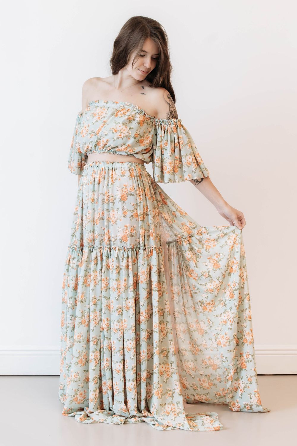 Woman wearing a floral off-shoulder maxi dress in mint green with peach flowers, holding out the tiered skirt.
