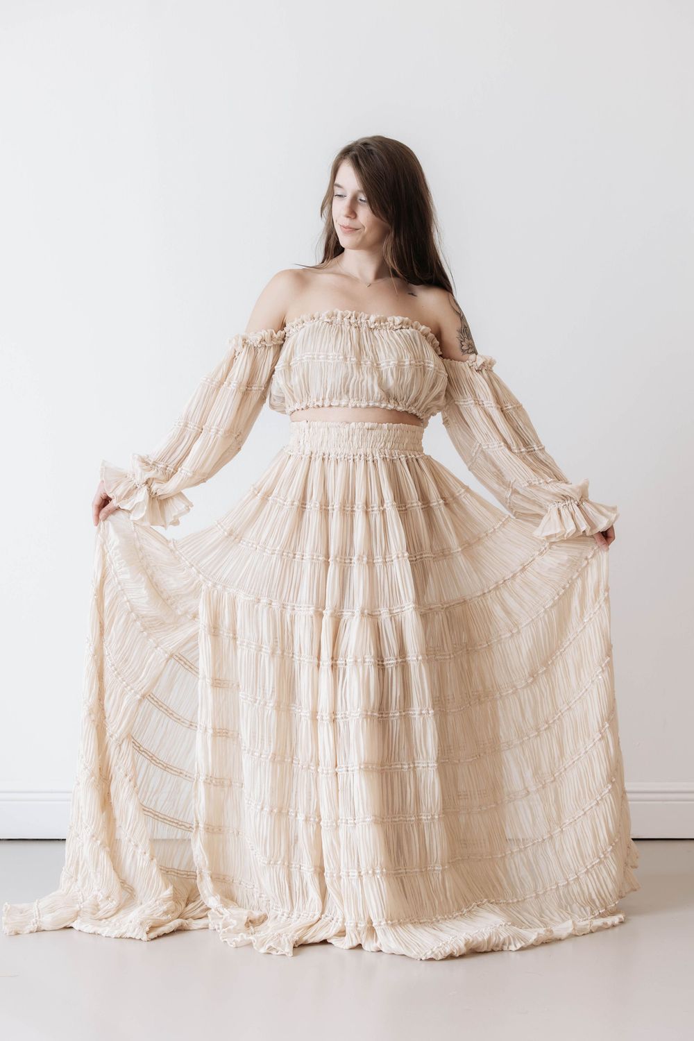 Woman in elegant off-shoulder cream chiffon tiered maxi dress with long sleeves, holding skirt wide in white studio.