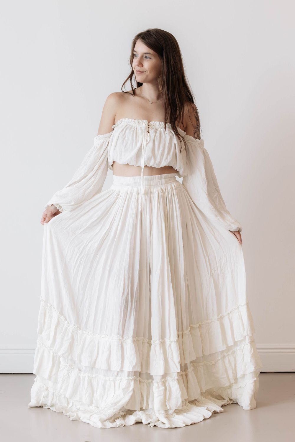 Woman wearing a white off-shoulder boho crop top and flowing maxi skirt set, posing against a white wall.