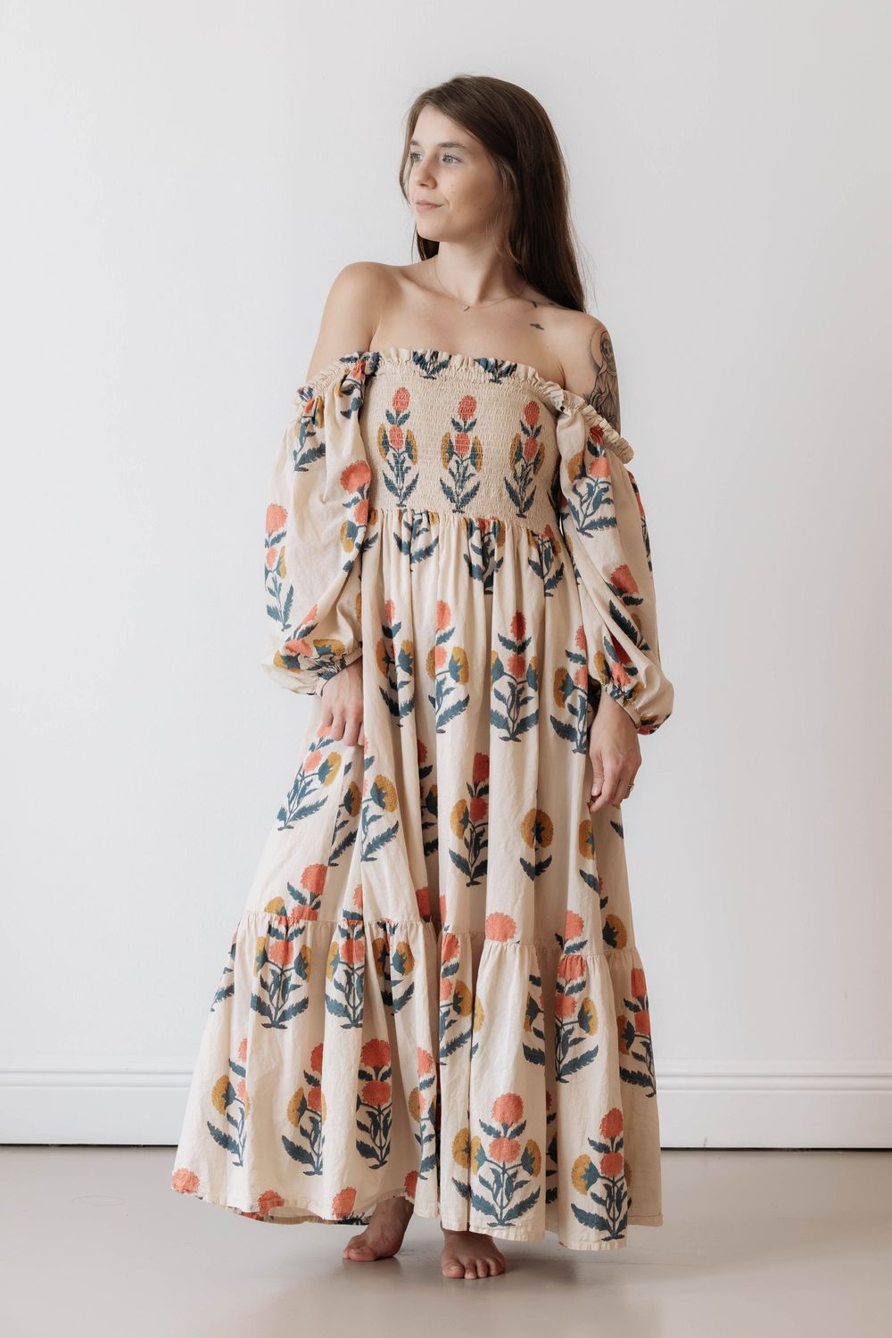 Woman wearing a floral off-shoulder maxi dress with puff sleeves and tiered skirt in cream with orange and green print.