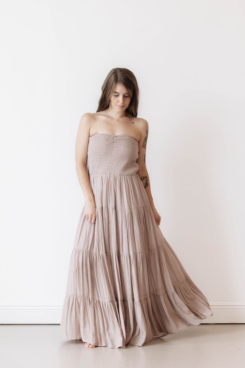 Woman in a flowing strapless tiered maxi dress in taupe, posing against a white wall.