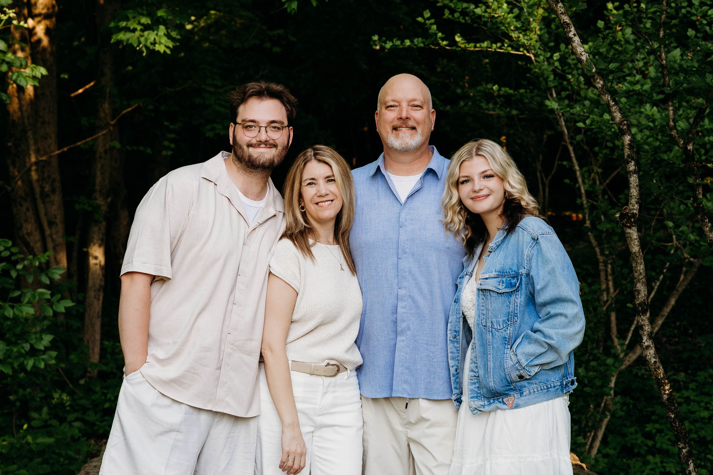 Family of four smiling together outdoors in front of lush green trees, dressed in casual white and blue outfits.