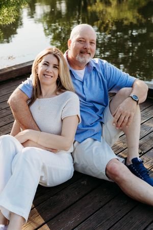 A smiling couple sitting together on a wooden dock by a serene lake on a sunny day.