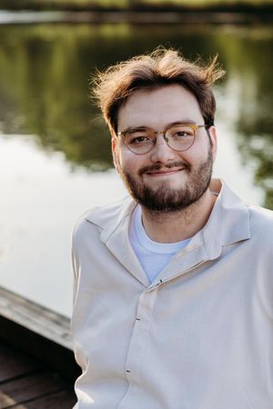 Young bearded man with glasses smiling outdoors near a reflective lake on a sunny day.