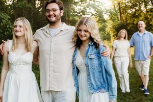 Family group smiling and walking outdoors in golden hour sunlight among lush green trees.