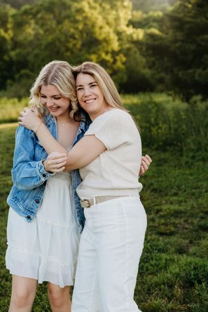 Two blonde women hugging and smiling outdoors in a green field, wearing white outfits and a denim jacket.