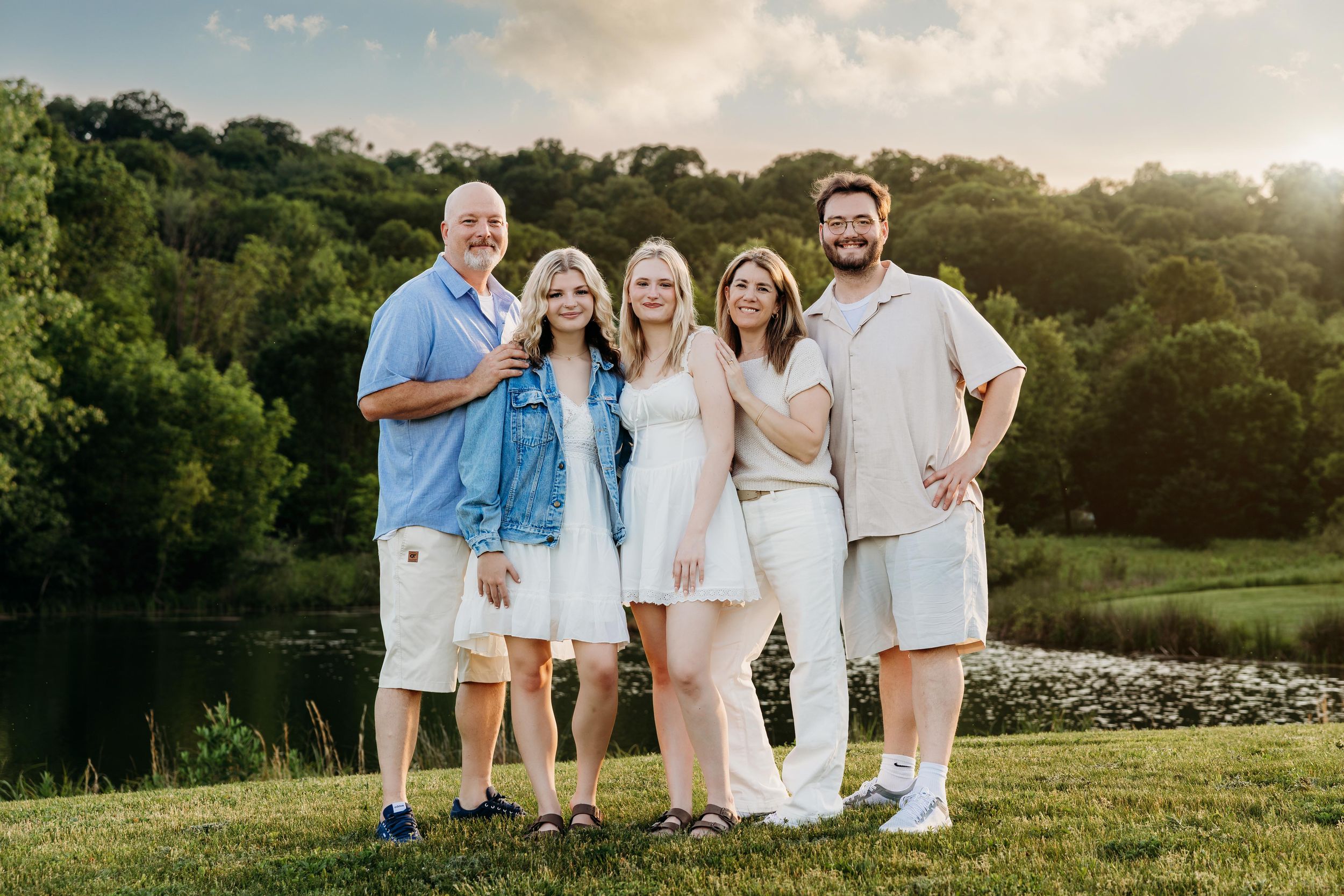 Family of five posing outdoors near a pond with lush green trees at golden hour, dressed in coordinated neutral outfits.