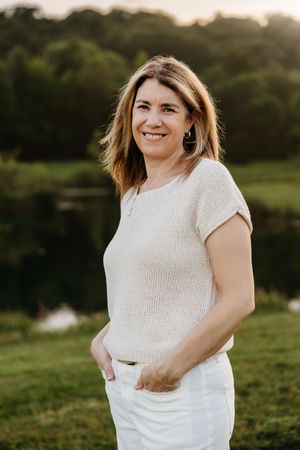Smiling woman in white knit top and pants standing outdoors near a pond with lush green trees at sunset.