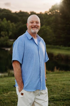 Smiling bald man with beard wearing blue shirt and khaki pants standing outdoors near a pond at sunset.