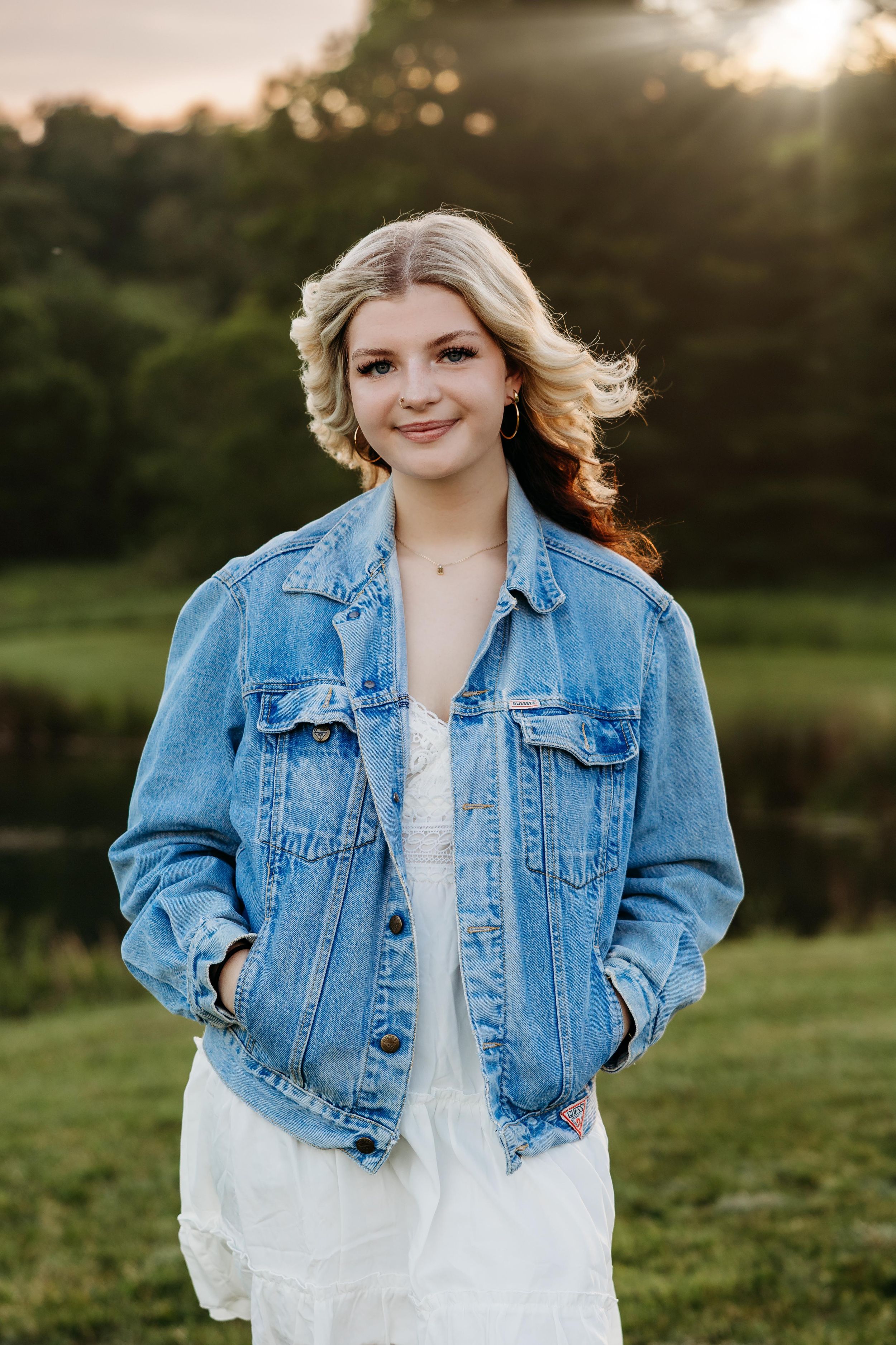 Young blonde woman in denim jacket and white dress smiling outdoors at golden hour.
