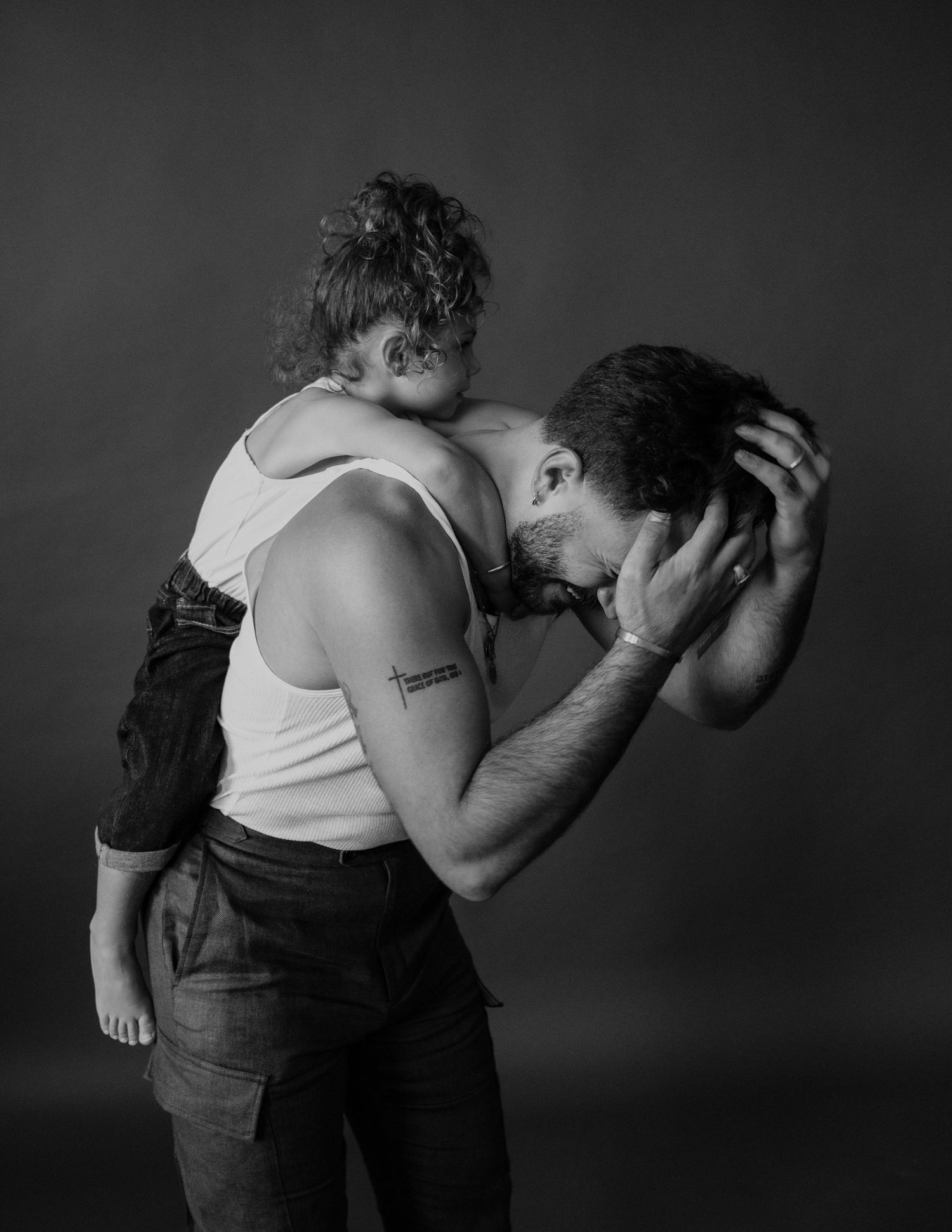 Black and white photo of a man carrying a child on his shoulder, bowing his head with hands clasped behind it.