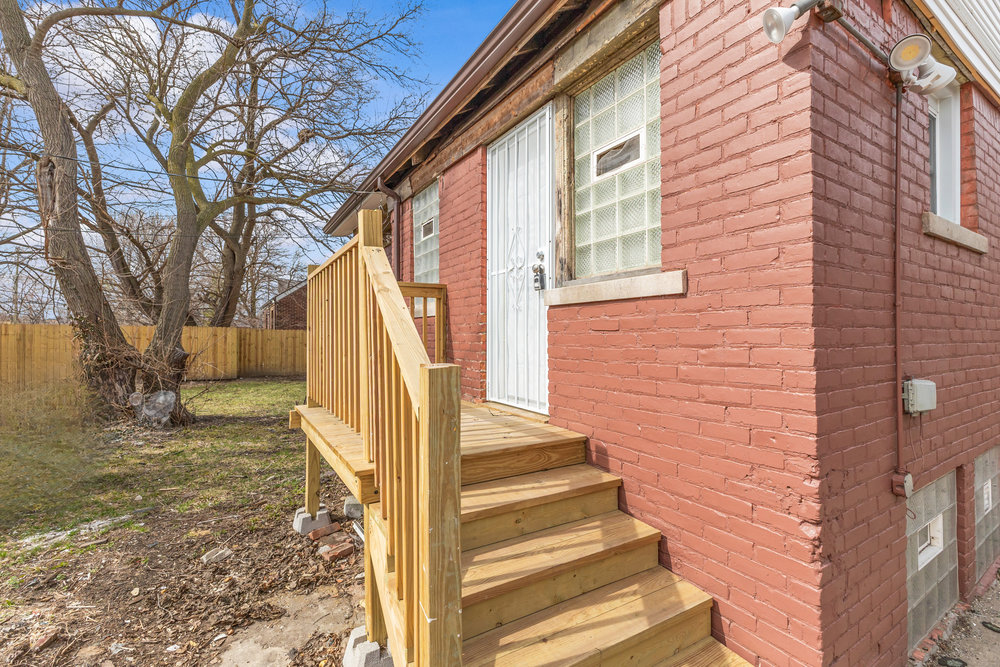 Red brick home exterior featuring a new wooden deck with stairs, white door, and glass block window in early spring. Detroit Michigan