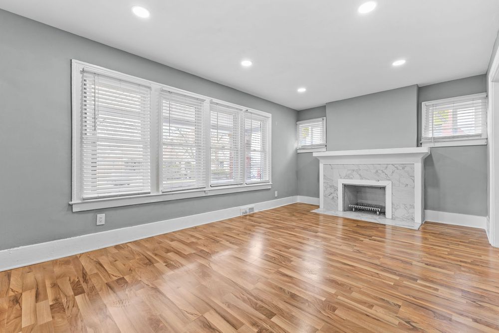 Bright empty living room with hardwood floors, white fireplace, gray walls, recessed lighting, and white shuttered windows. Detroit Michigan