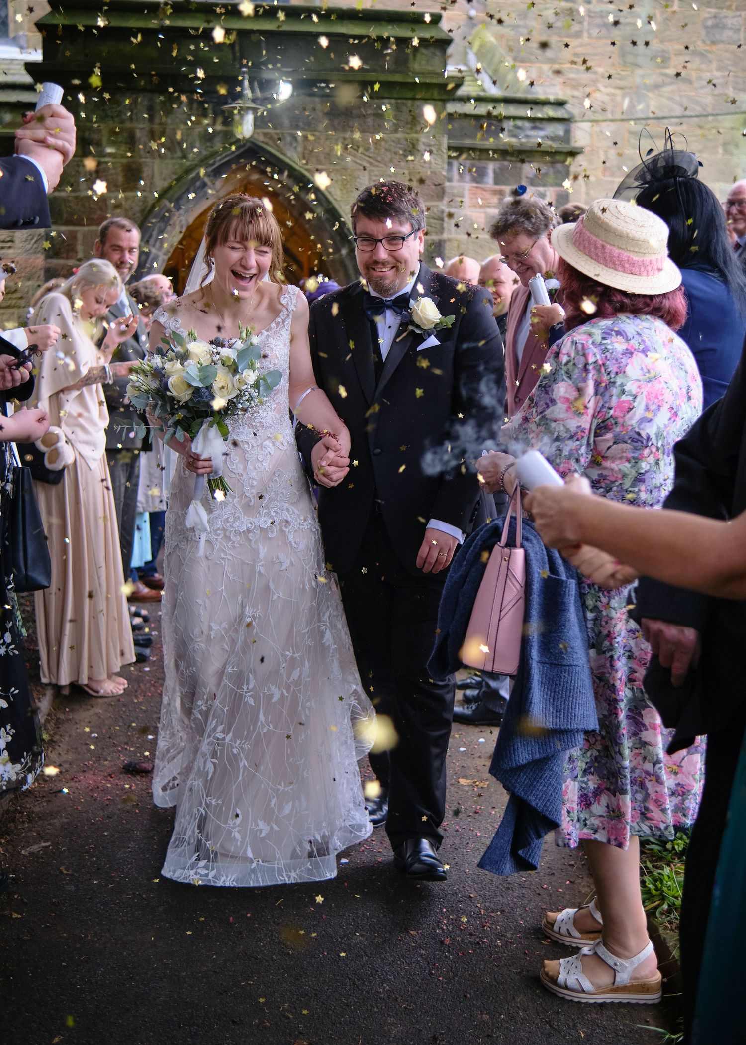 Bride and groom walk through confetti shower after wedding ceremony, surrounded by cheering guests outside a stone church.