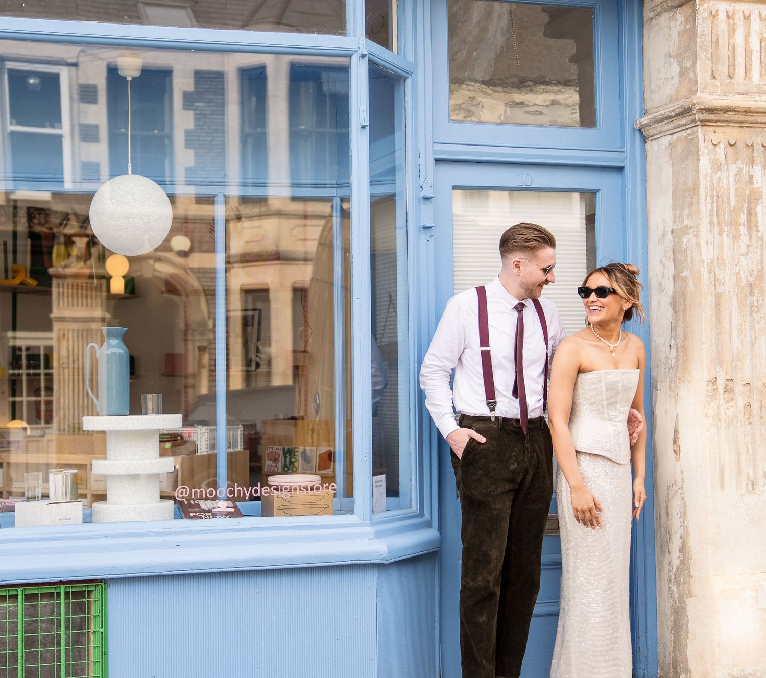 A bride and groom pose outside a blue-framed shop front, smiling at each other in elegant wedding attire.