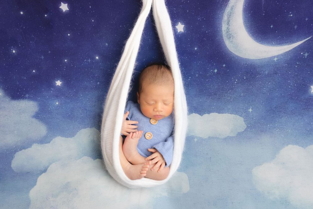 Baby sleeps peacefully in white hammock against dreamy night sky backdrop with crescent moon and stars.