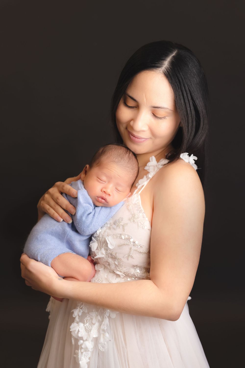 A tender moment captured as a mother in a white lace dress holds her sleeping newborn baby wearing soft blue clothing.