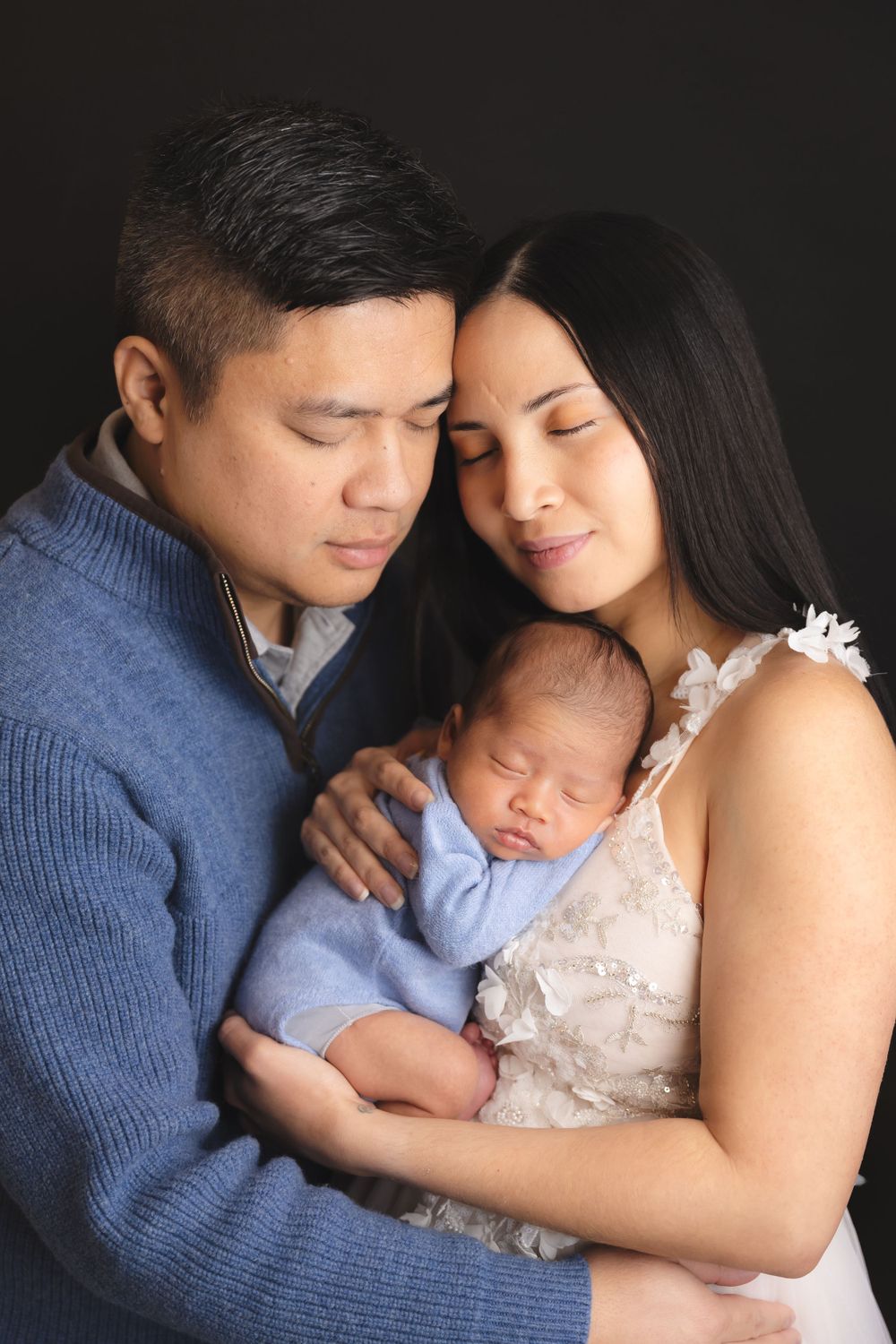 A couple in blue and white hold their sleeping newborn baby against a dark background in an intimate portrait.