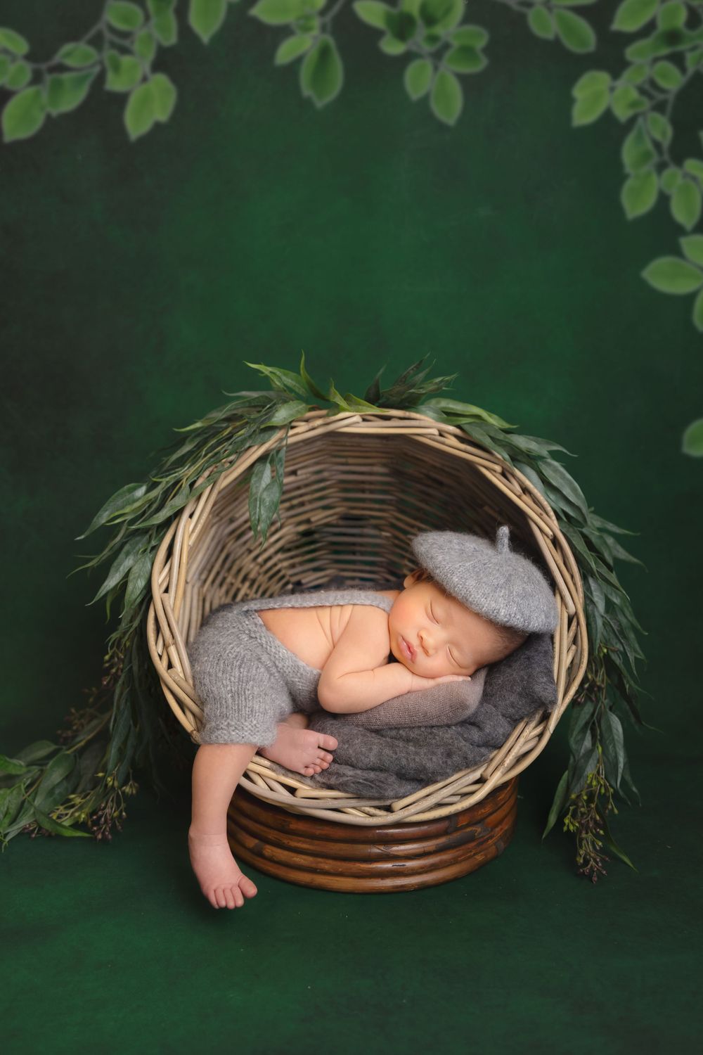 A sleeping baby nestled in a woven basket with gray knit clothing against a dark green backdrop with leafy foliage.