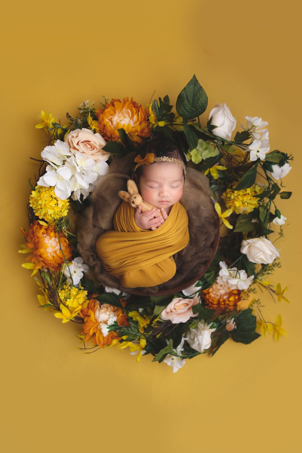 A sleeping newborn wrapped in mustard yellow fabric lies in a wooden bowl surrounded by colorful autumn flowers on yellow backdrop.