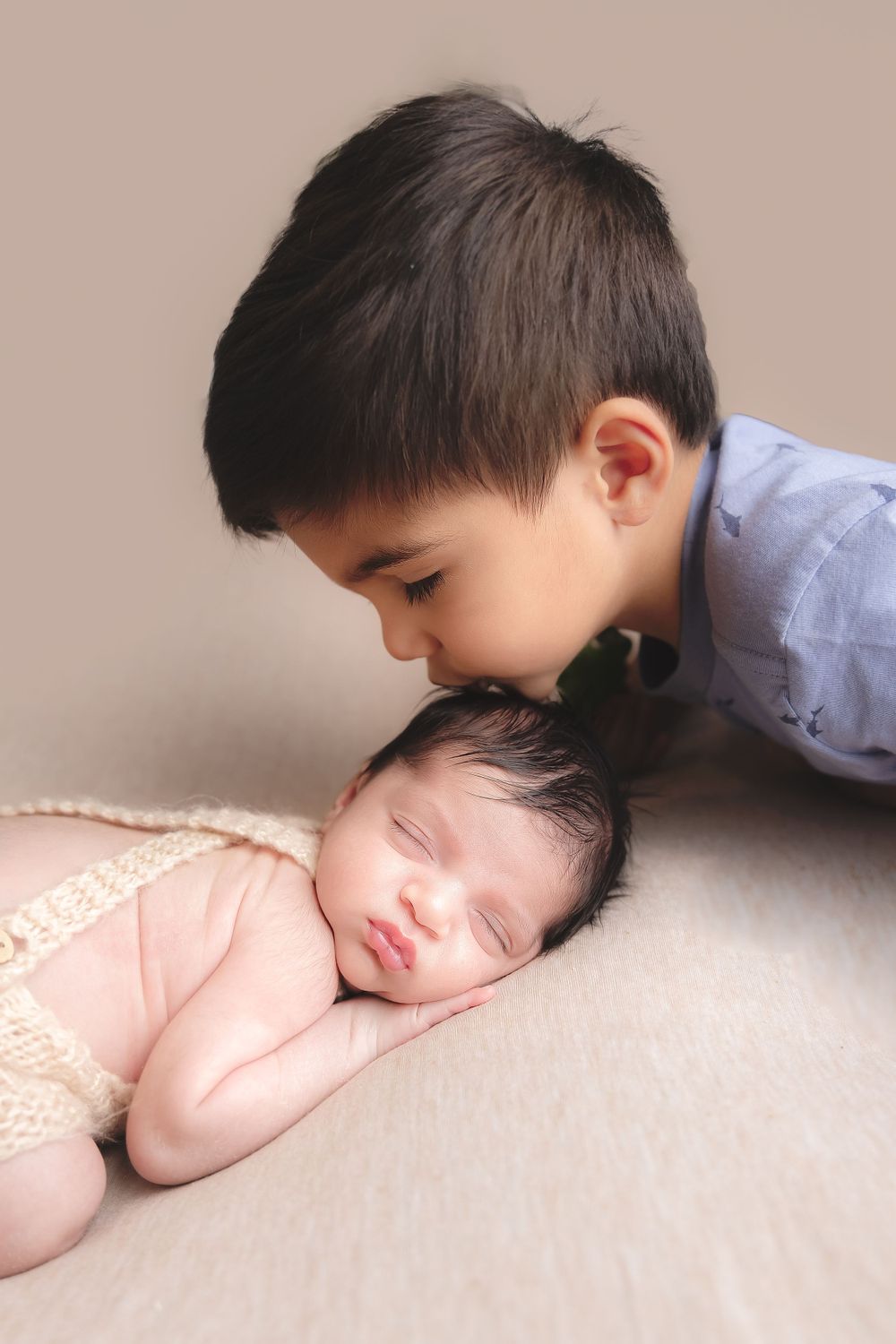 A young boy in a blue shirt gently kisses his sleeping newborn sibling who wears a lacy outfit in newborn photography session Sammamish WA