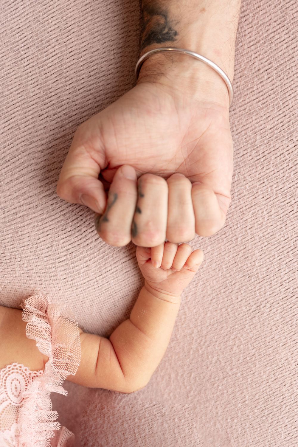 A small baby's hand gently holds an adult's hand wearing a silver bracelet against a soft pink background. newborn photography session Samma
