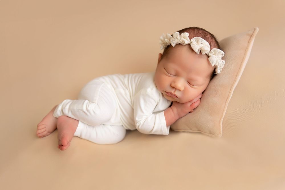 Sleeping newborn wearing white outfit and floral headband rests peacefully on beige cushion during professional photoshoot.