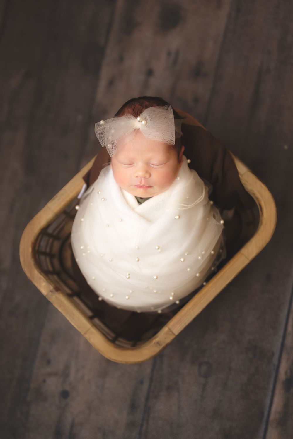 Newborn baby swaddled in white fabric with a bow headband sleeping peacefully in a wooden basket on dark wooden floor.