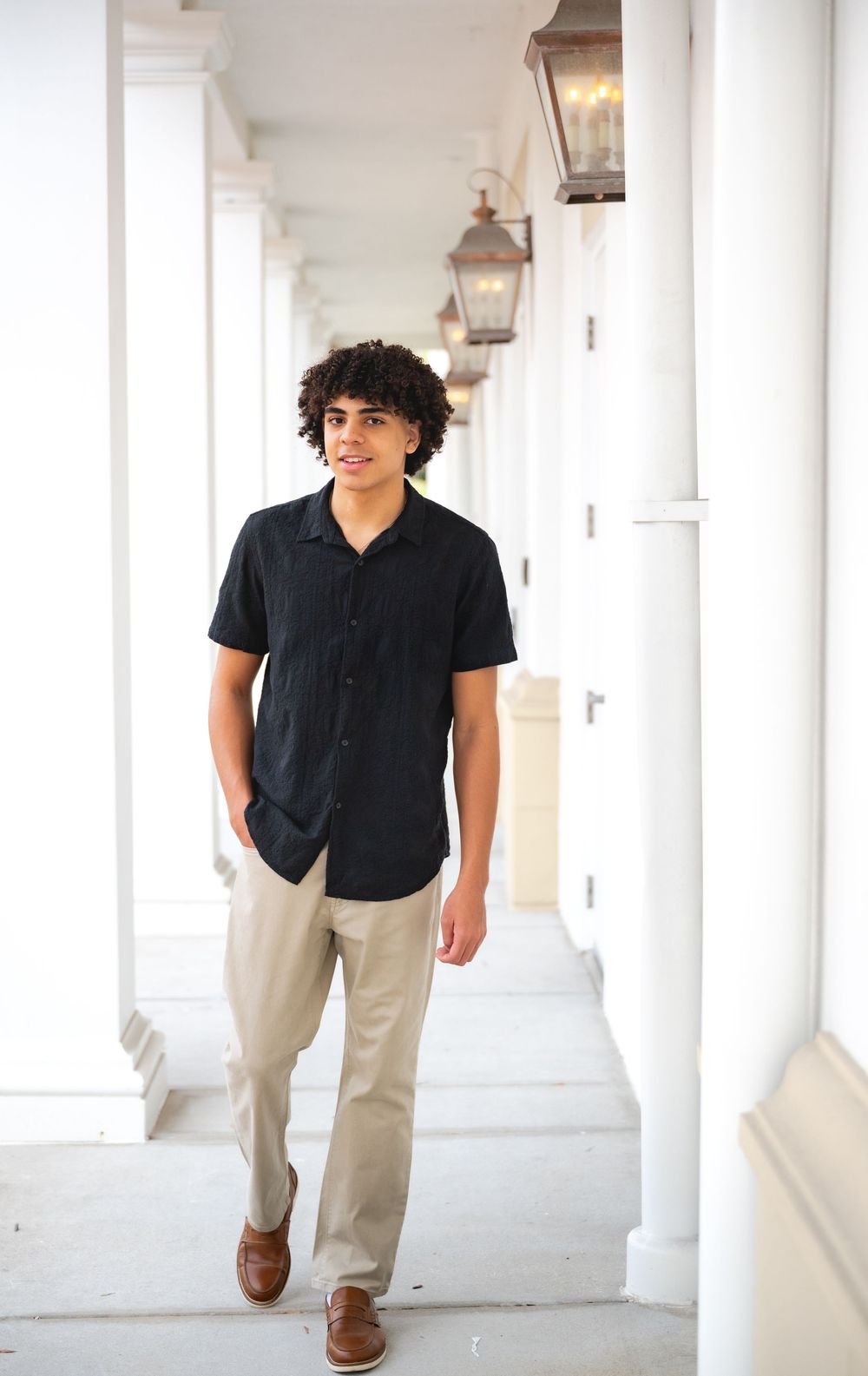 Young man with curly hair walks confidently in black shirt and khaki pants along a white columned hallway.