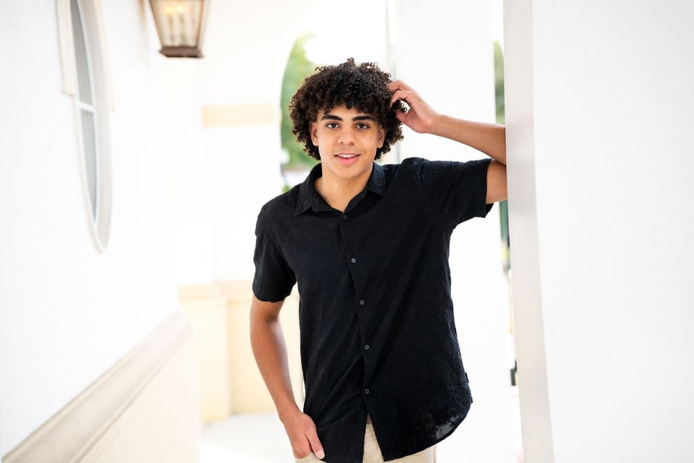 Young man in black shirt leaning against white pillar, smiling outdoors in bright natural light.