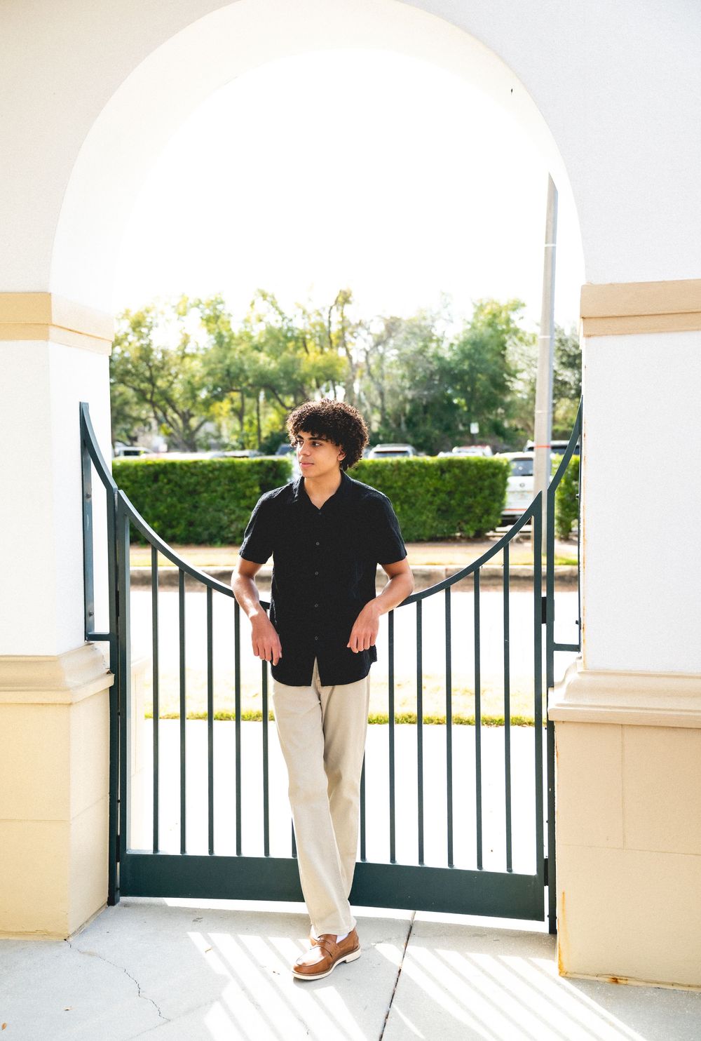 Young man in black shirt and khaki pants standing by an ornate iron gate under a white archway.