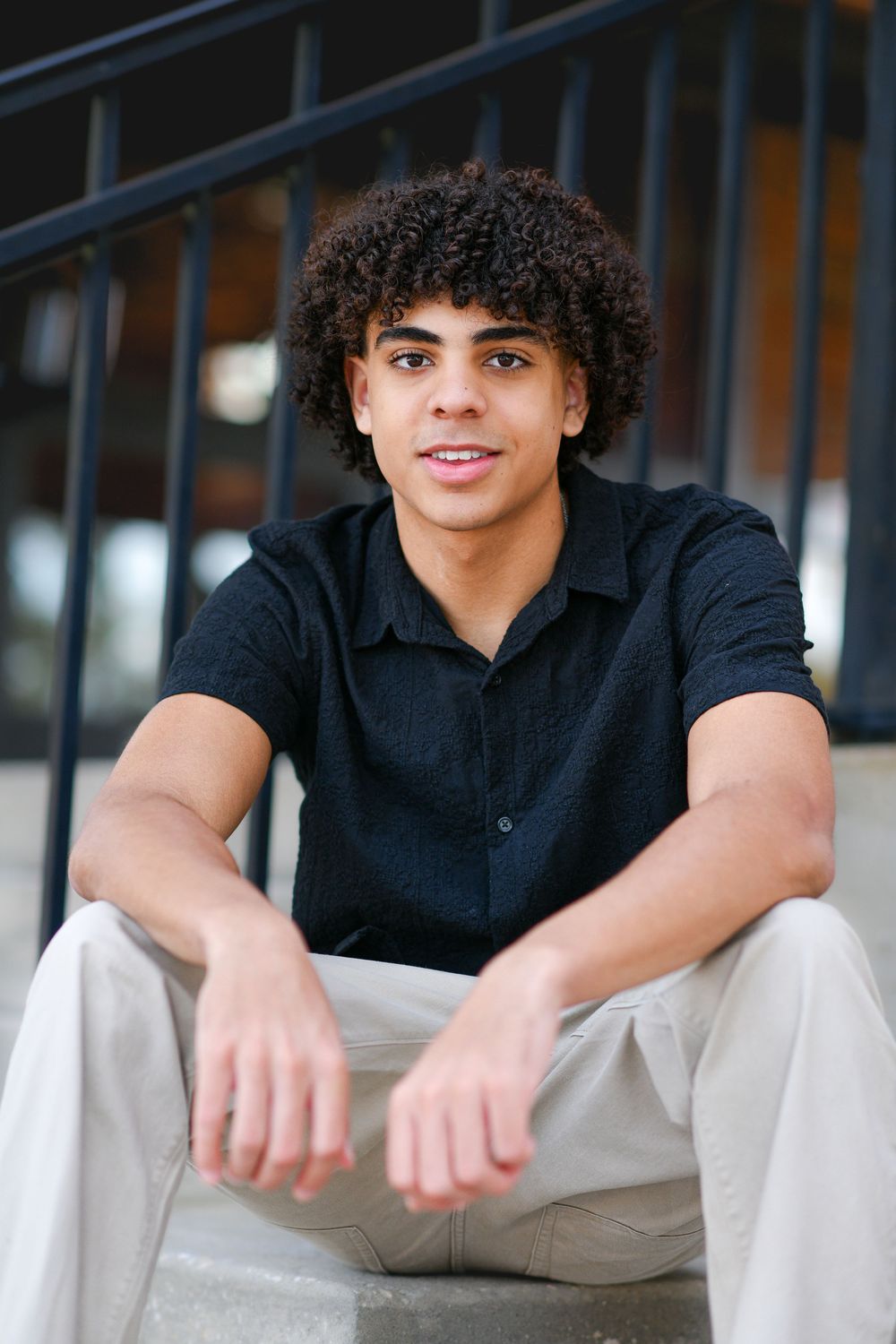 Young man with curly hair sitting on steps wearing a black shirt and khaki pants, smiling at camera.