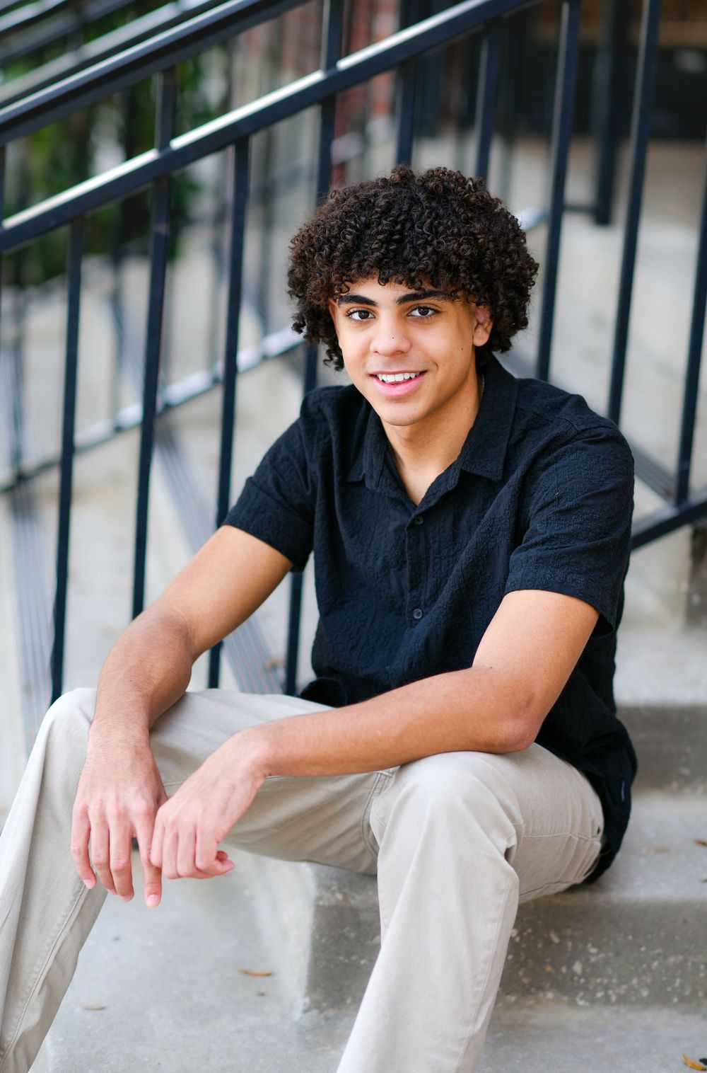 A smiling young man with curly hair sits on outdoor steps wearing a black shirt and khaki pants.