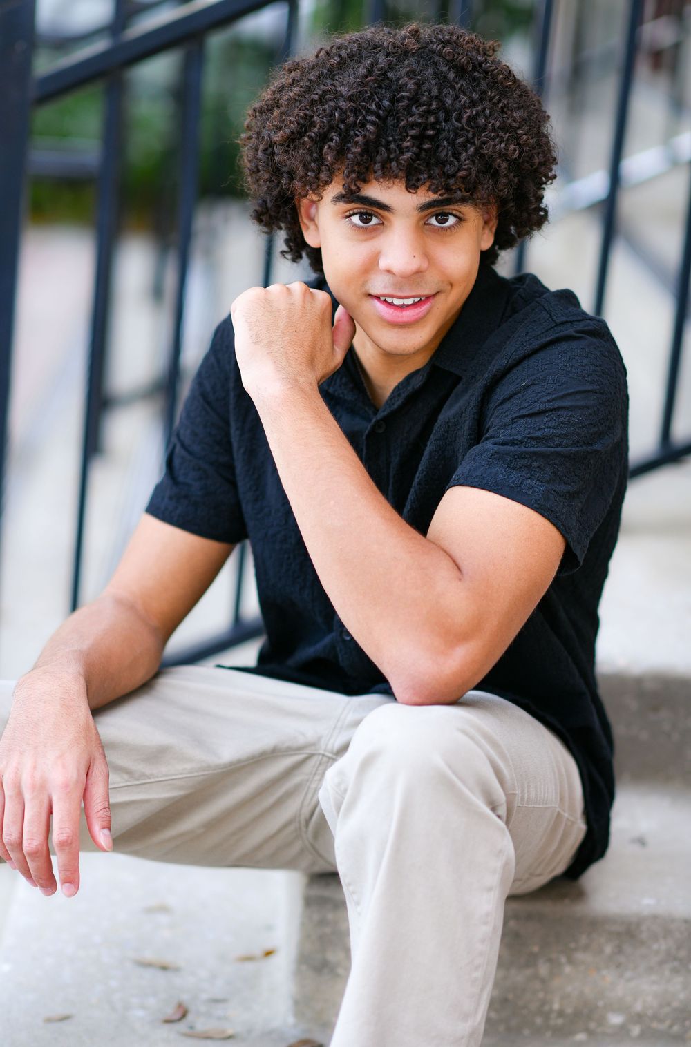 Young man with curly hair sitting on steps, smiling in black polo shirt and khaki pants.