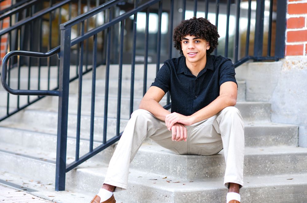 Young man with curly hair sitting on outdoor steps wearing a black shirt and khaki pants.
