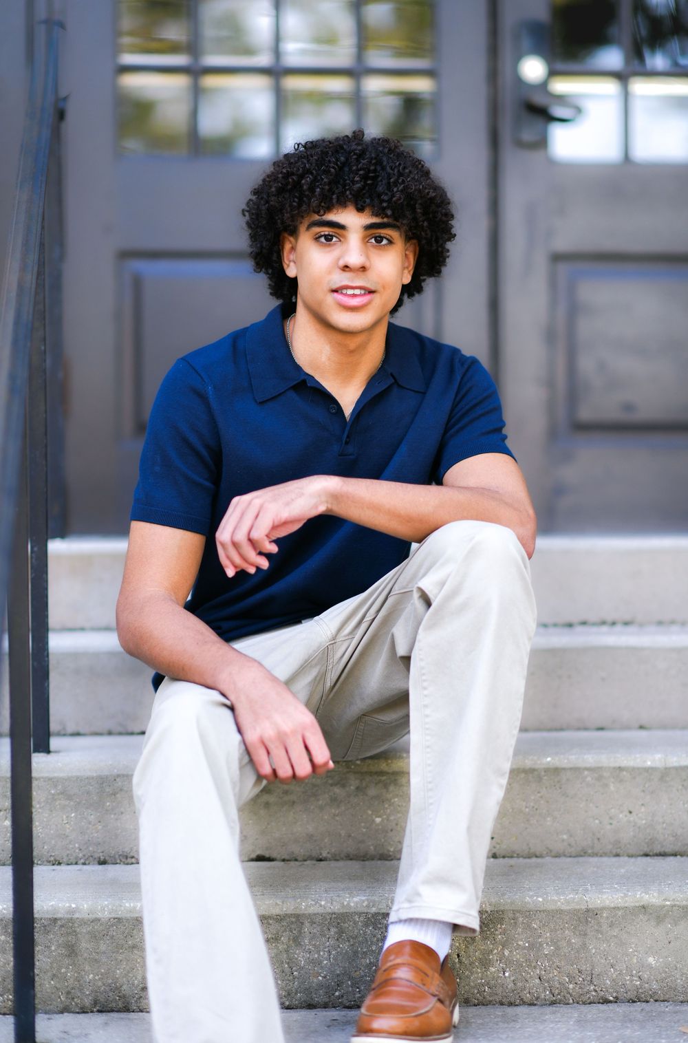 Young man with curly hair sitting on steps wearing a navy polo shirt and khaki pants with brown loafers.