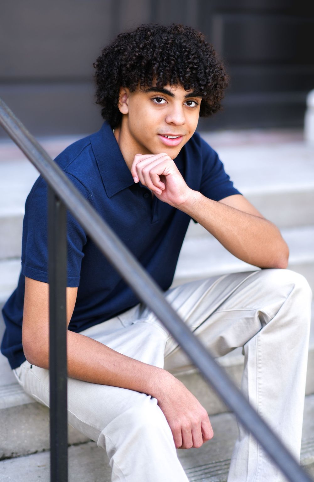 Teen boy with curly hair sitting on steps in navy polo shirt and khaki pants, resting chin on hand.