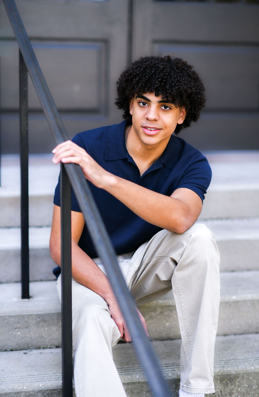 Young man with curly hair sits on steps, wearing navy polo and khaki pants, smiling at camera.