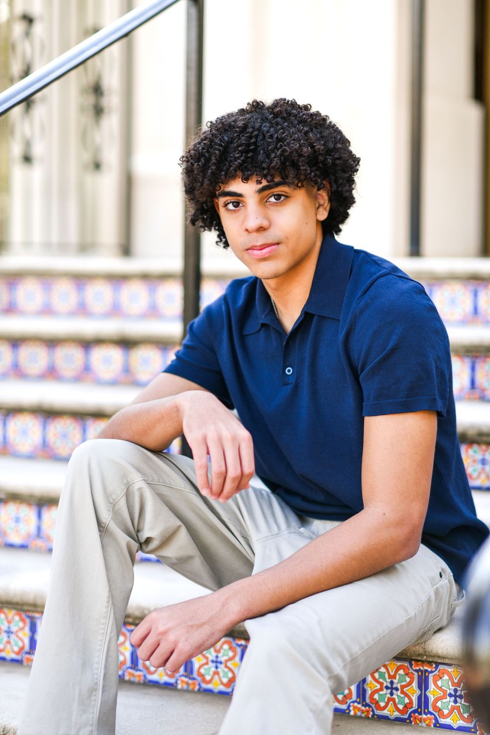 Teen boy with curly hair sitting on decorative tiled steps in navy polo and khaki pants.