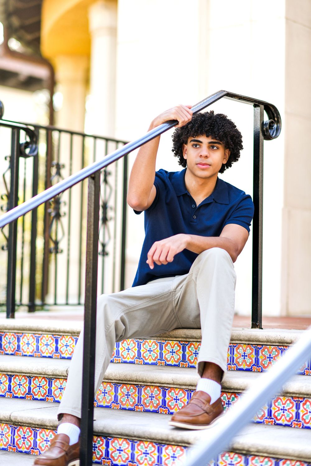 Young man with curly hair sits on colorful tiled steps, wearing navy polo and khakis, leaning on black iron railing.