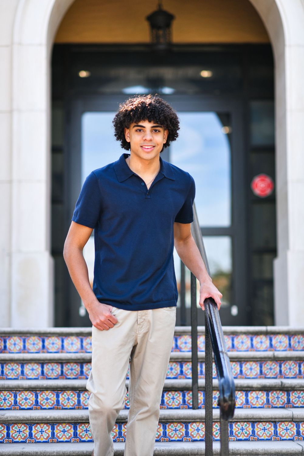Smiling teen with curly hair stands on tiled steps in navy polo and khakis outside building.