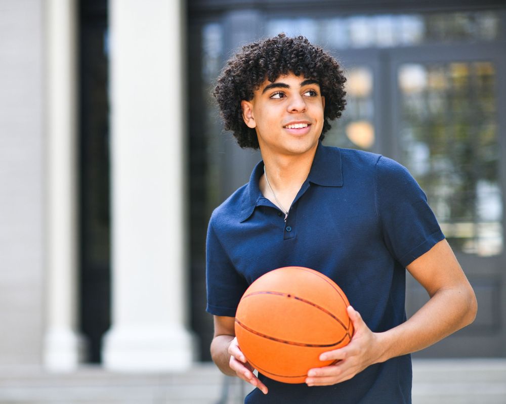 Young man with curly hair holding a basketball outdoors, wearing a navy polo shirt, looking away confidently.