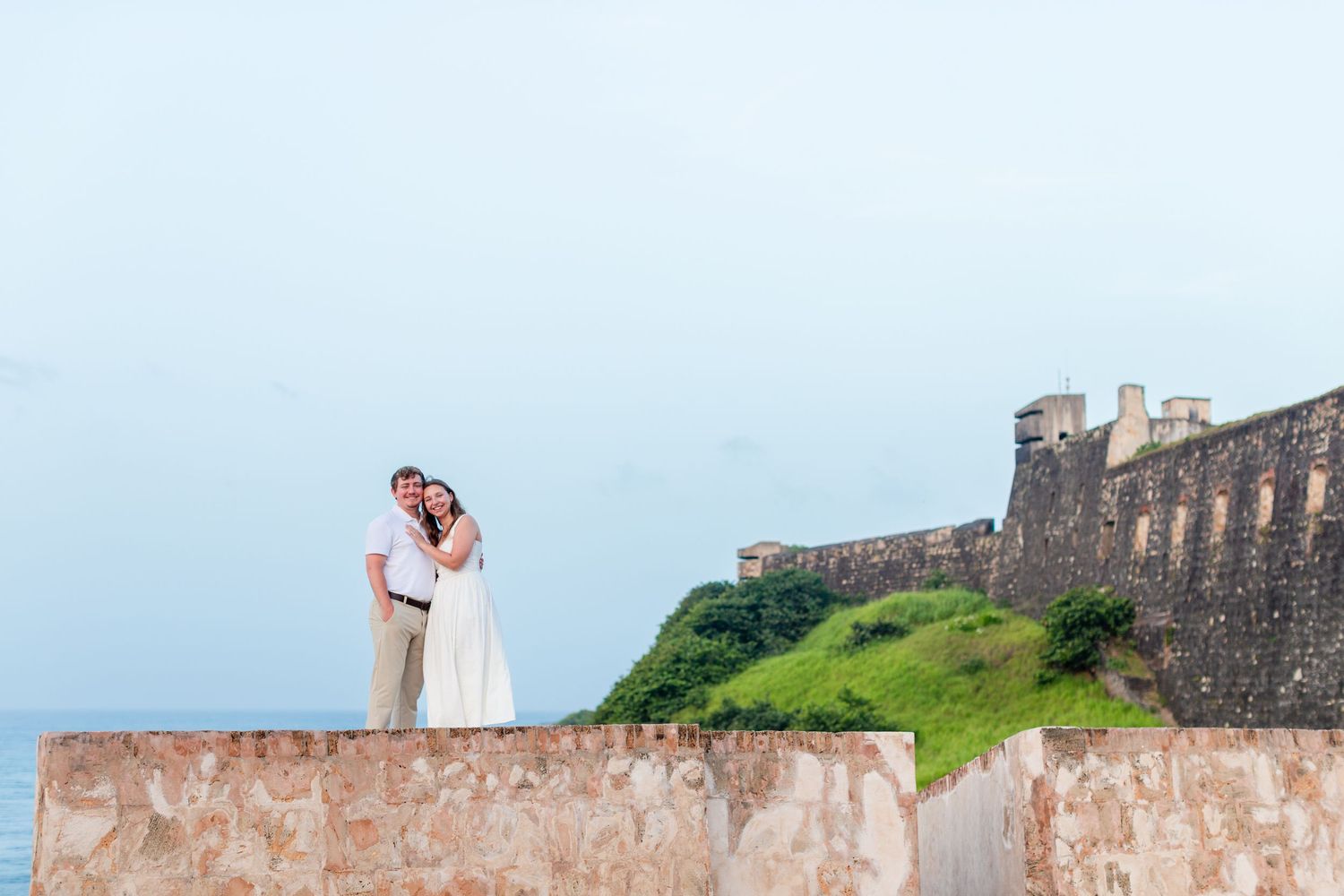 Couple standing on fort walls with Castillo San Cristóbal in the background. Engagement photo session in Old San Juan, Puerto Rico.